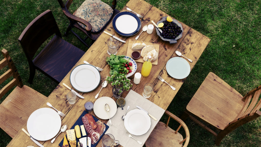 Eettafel in de tuin gedekt voor zes personen met een mix van houten stoelen en een verscheidenheid aan gerechten, inclusief brood, vleeswaren en verse groenten.