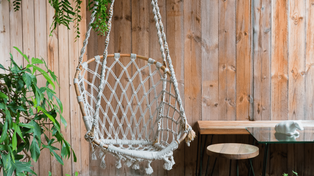 Decoratieve hangstoel met macramé patroon en houten omlijsting tegen een houten wand, naast groene planten en een minimalistische tafel.