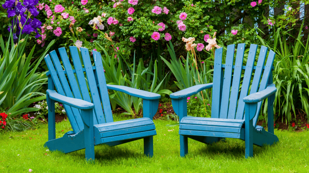 Twee blauwe Adirondack-stoelen in een groene tuin met kleurrijke bloemen op de achtergrond.