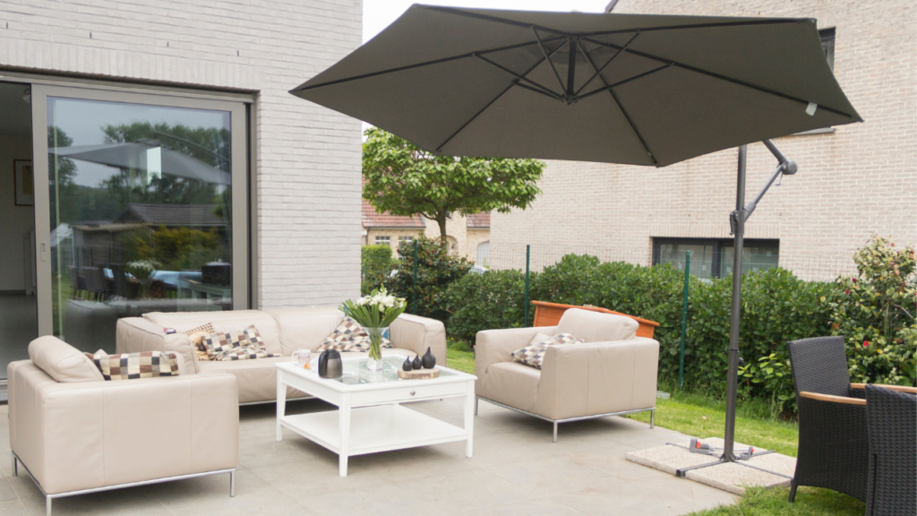 Stijlvolle tuinset met beige lederen banken, witte salontafel en een grote zwarte parasol op een betegeld terras naast een modern huis.